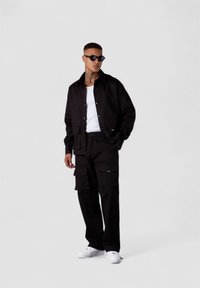 Schwarze Baumwolljacke mit Knopfverschluss, kombiniert mit passenden Cargo-Hosen mit Taschen. Das Model trägt ein weißes Tanktop und Sneakers, accessoriert mit schwarzen Sonnenbrillen.