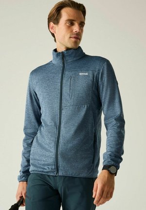 Mann trägt eine blaue Jacke mit Reißverschluss und hohem Kragen sowie eine schwarze Smartwatch, blickt zur Seite und hält einen braunen Gegenstand in der linken Hand.