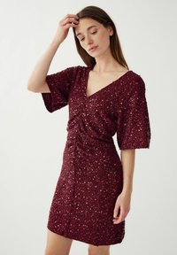 Vyno raudonos spalvos sequin suknelė su V formos iškirpte, trumpomis rankovėmis ir susiraukusiais detalėmis ties juosmeniu, sukurianti prigludusį siluetą.