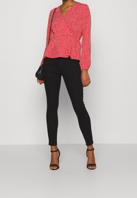 Blusa rossa con pois bianchi, design avvolgente e maniche lunghe, abbinata a pantaloni neri aderenti e tacchi neri con cinturini, tenendo una borsa nera.
