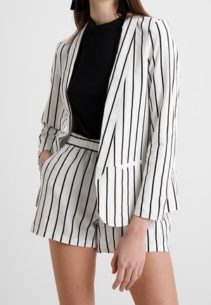 Kvinde iført en hvid blazer og shorts med lodrette sorte striber over en sort top, hånd i lommen, mod en ensfarvet baggrund.