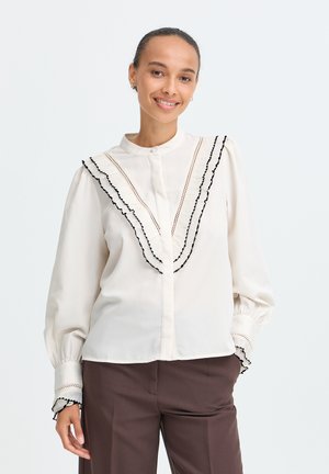 Frau lächelt, trägt eine weiße Bluse mit schwarzem Rüschenbesatz und braune Hose, steht vor einem schlichten hellen Hintergrund.