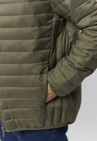 Geacă matlasată verde măsliniu cu textură cusută orizontal, prevăzută cu buzunar cu fermoar și manșete elastice, vazută din lateral și din spate.