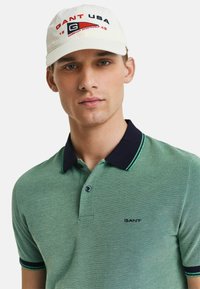 Camisa polo verde texturizada con cuello y puños azul marino oscuro, que presenta un pequeño logotipo bordado. La gorra tiene una base crema con acentos rojos y azules.