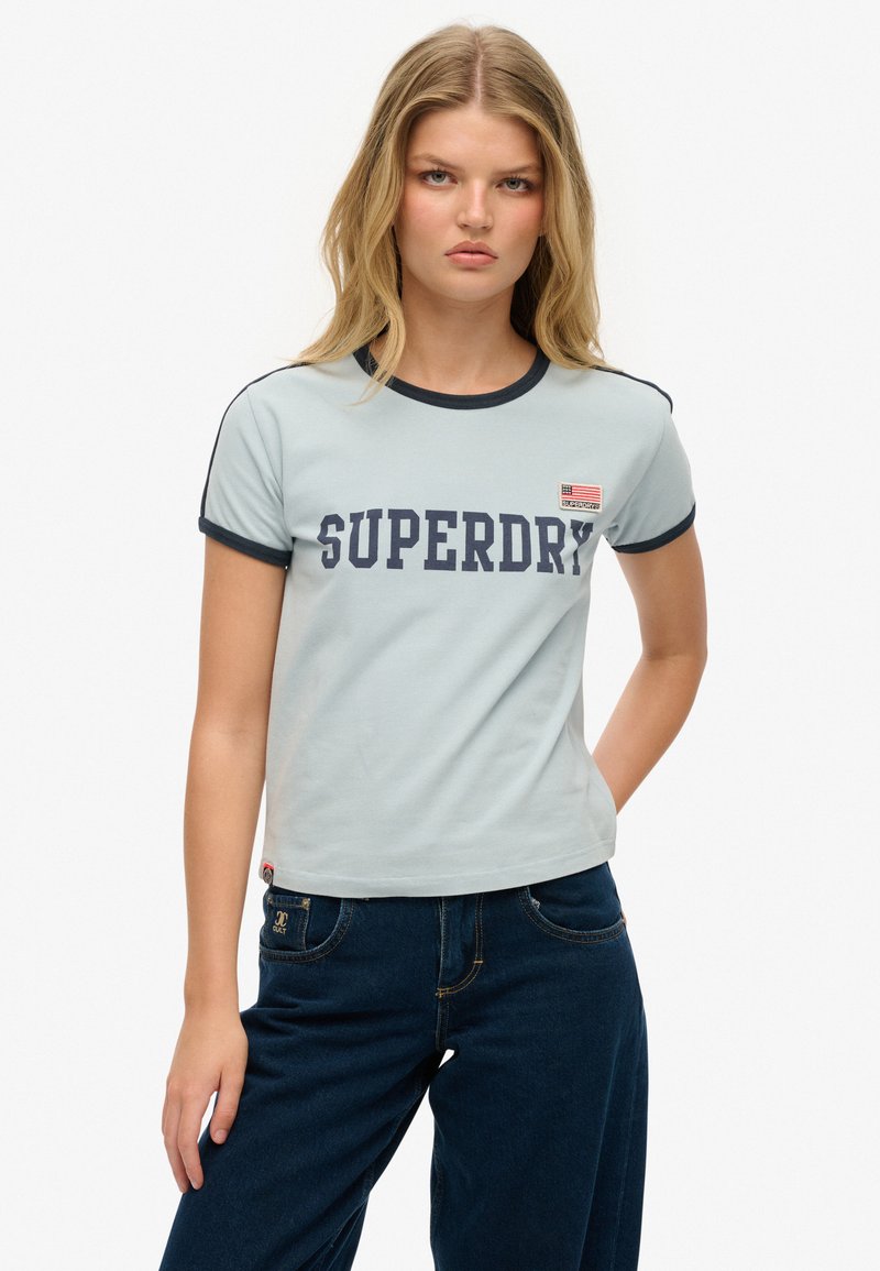Hellblaues Baumwoll-T-Shirt mit marineblauen Abschlüssen und dem Schriftzug "SUPERDRY" in fetten Buchstaben. Auf der Brust befindet sich ein kleines patch in Form einer Flagge.