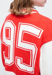 Červený a biely pletený sveter s veľkým číslom "95" na chrbte. Tkanina má textúrovaný vzor a kontrastný rebrovaný golier.