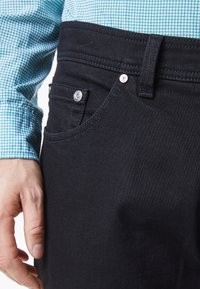 Hombre que lleva jeans de mezclilla negros con un bolsillo frontal visible y un lazo para el cinturón, combinado con una camisa de manga azul con un patrón, y con la mano descansando cerca de la cadera.