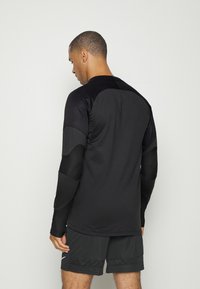 Schwarzes Langarm-Sportshirt mit strukturierten Ellenbogenpatches und Schulterakzenten, aus einem glatten, dehnbaren Material gefertigt. Kombiniert mit dunklen Shorts.