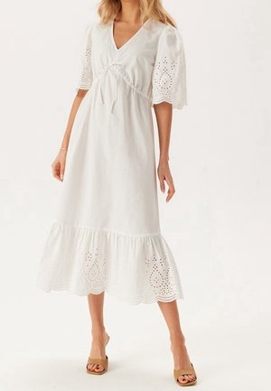 Mujer con un vestido midi blanco con mangas abullonadas y bajo de encaje con ojales, parada con sandalias marrones de tacón ancho y punta abierta sobre un fondo liso.