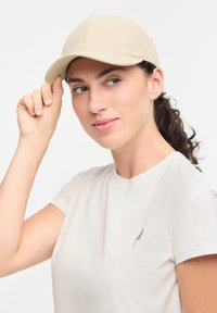 Beige pet met een ronde kroon en gebogen rand, voorzien van een klein geborduurd boompje aan de zijkant. Model draagt een lichtgekleurde T-shirt.
