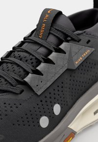 Sneakers de trail Nike noires avec tige perforée, superpositions texturées, accents gris et marquage orange. Comprend des lacets plats et un col rembourré.