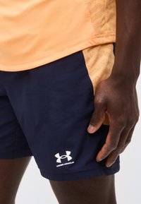 Άτομο με σκουρόχρωμο δέρμα που φοράει σκούρα μπλε σορτς Under Armour και ανοιχτό πορτοκαλί πουκάμισο, με το χέρι να ακουμπά κοντά στη ραφή των σορτς.