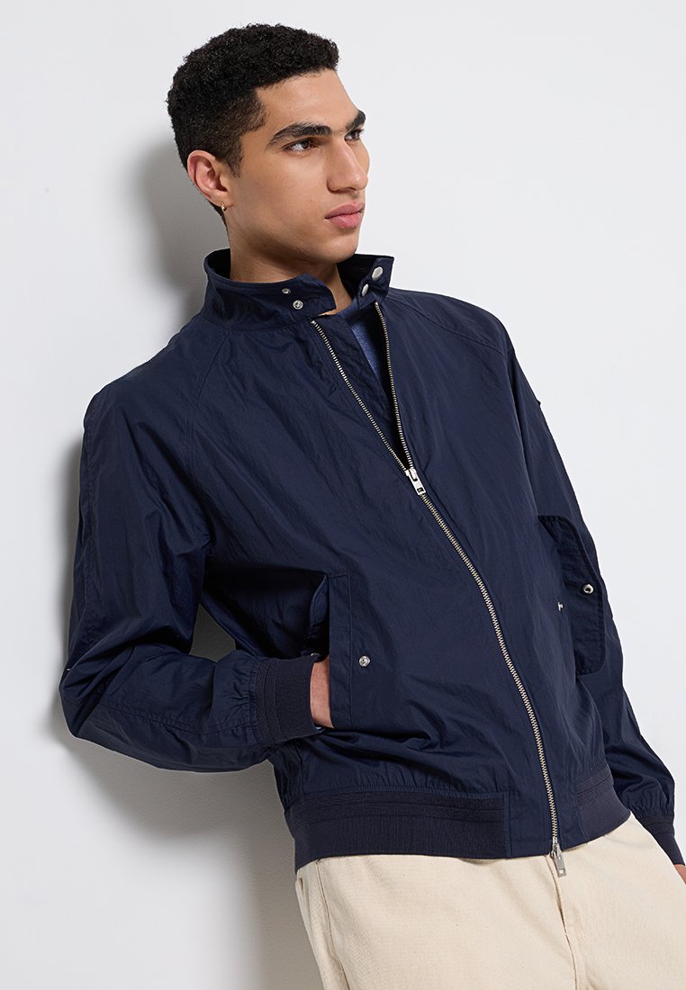 Marineblaue leichte Bomberjacke mit Reißverschluss vorne, hohem Kragen und gerippten Bündchen. Glatte Textur mit seitlichen Taschen und metallischen Beschlägen.