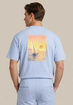 Man with brown hair wears light blue shirt and pants; shirt back shows sailboat on water with a sunset background.