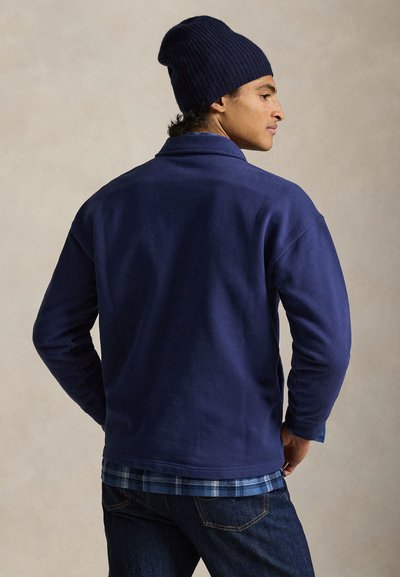 Άνδρας που φοράει ναυτικό σκουφάκι, ναυτικό φλις μπουφάν, μπλε πουκάμισο με καρό σχέδιο και σκούρα τζιν, στέκεται πλευρικά μπροστά σε απλό φόντο.