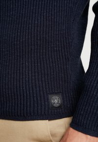 Maglione a coste blu navy con finitura testurizzata e una toppa con logo quadrato sull'orlo. Caratterizzato da maniche lunghe e un design aderente.