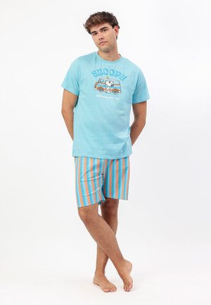 Joven de pie descalzo, vistiendo una camiseta ligera azul claro con Snoopy y pantalones cortos a rayas verticales naranjas y azules sobre fondo blanco.