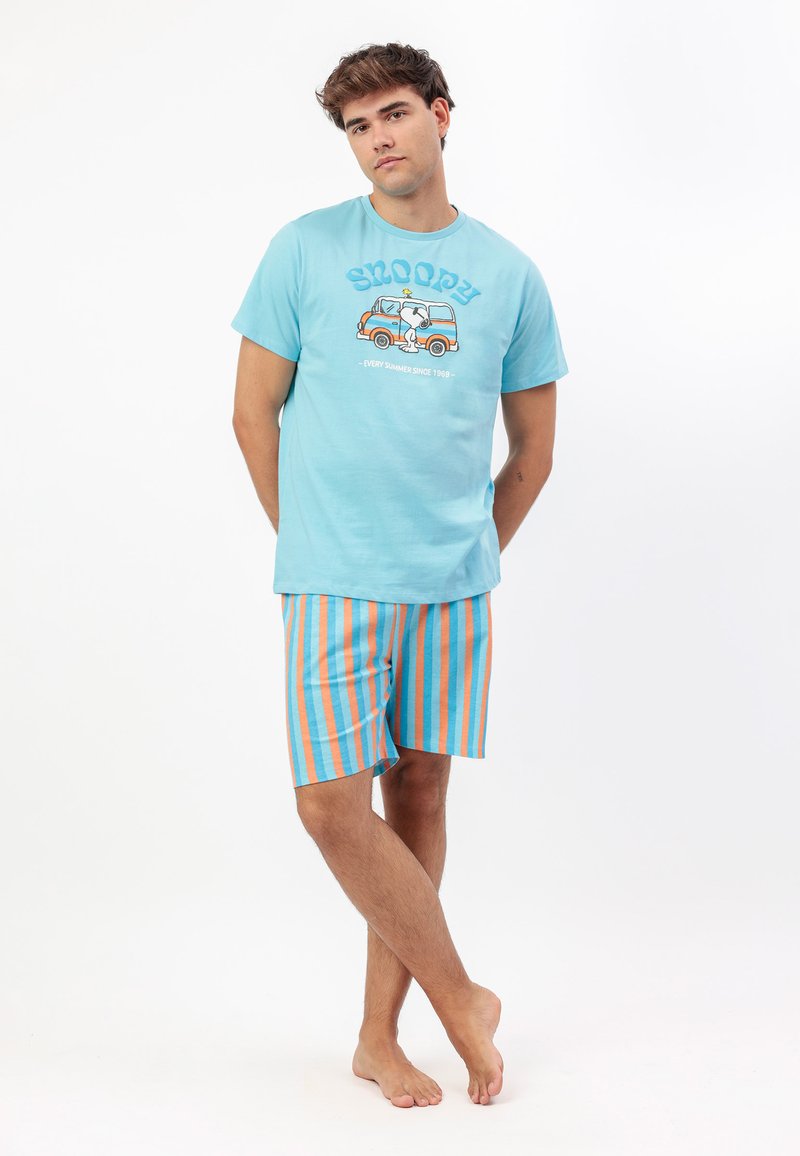 Joven de pie descalzo, vistiendo una camiseta ligera azul claro con Snoopy y pantalones cortos a rayas verticales naranjas y azules sobre fondo blanco.