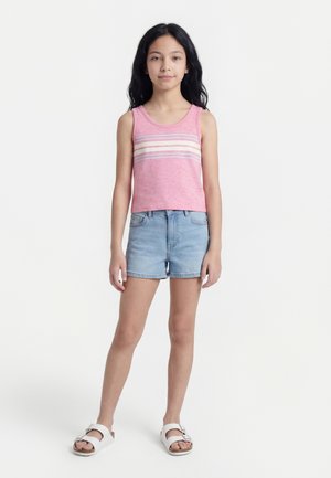 Κορίτσι που στέκεται φορώντας ροζ αμάνικη μπλούζα με ρίγες, ανοιχτόχρωμα τζιν σορτς και λευκά σανδάλια με αγκράφες, σε λευκό φόντο.