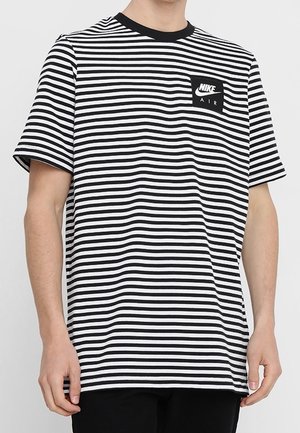 Homme portant un T-shirt Nike Air à rayures horizontales noires et blanches, manches courtes, avec un col noir, debout devant un fond uni.