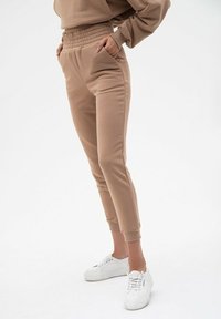 Beżowe dresy z wysokim, elastycznym pasem, bocznymi kieszeniami i zwężanymi mankietami. Noszone z białymi trampkami na jednolitym tle.