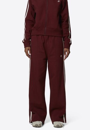 Persona che indossa pantaloni della tuta bordeaux e giacca con strisce bianche ai lati e scarpe da ginnastica grigie, in piedi contro uno sfondo semplice.