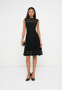 Vestido preto sem mangas com um corpete texturizado e saia rodada, apresentando riscas horizontais transparentes na gola e na barra. Usado com sapatos de salto pretos.