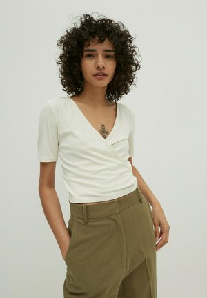 Weißes geripptes Oberteil mit kurzen Ärmeln und tiefem V-Ausschnitt, kombiniert mit hoch taillierten olivgrünen Hosen. Einfaches Design und glatte Textur.