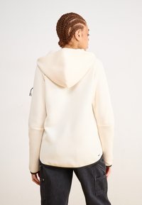 Sweatshirt claro de creme com cordão ajustável, textura macia e bolsos canguru, combinado com calças cargo escuras, visto de costas.