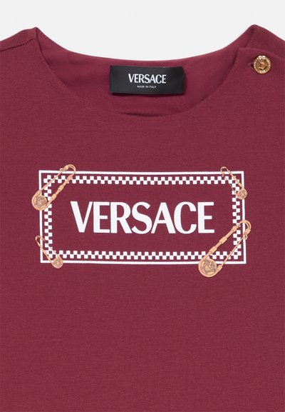 Vīna sarkana kokvilnas t-krekls ar baltu "VERSACE" logo, ko ierobežo rūtainā raksta malas. Zelta drošības adatas akcentē dizainu tuvu kakla apdarei.