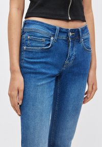 Kék farmernadrág karcsúsított szabással, közép magas derékkal, öt zsebbel és kontrasztos varrással. A farmer anyaga sima textúrájú.