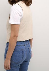 Gilet corto beige realizzato in materiale liscio, abbinato a una maglietta bianca e jeans di denim blu. La vista posteriore mostra un design aderente e tasche posteriori.