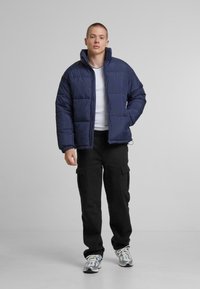 Marineblå pufferjakke med høj krave, der har en quiltet design, frontlynlås og elastiske cuff. Kåret med sorte cargo-bukser og sneakers.