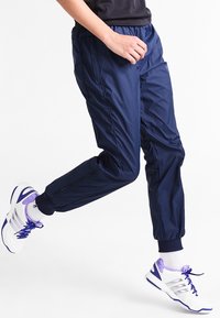 Marineblaue Jogginghose mit elastischem Bund und Reißverschlüssen an den Manschetten. Hergestellt aus glattem, leichtem Material. Getragen mit weißen und lila Sportschuhen.