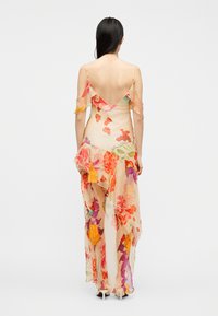 Femme aux longs cheveux foncés portant une robe maxi transparente à fleurs et épaules dénudées avec des volants, debout devant un fond clair uni.
