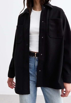 Woman wearing a black oversized button-up shirt with two chest pockets, white fitted T-shirt, blue jeans, and a black belt.