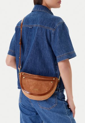 Persona vistiendo una camisa de mezclilla azul y jeans, llevando una bolsa cruzada de cuero marrón en forma de media luna con herrajes dorados en la espalda.