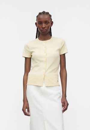 Noor inimene punutud juustega, kellel on seljas beež lühikeste varrukatega nööpidega topp ja valge seelik, seisab ühevärvilise heleda tausta ees.