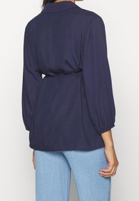 Mörkblå topp med krage, långa ballongsärmar och midjeband, tillverkad av ett slitstarkt tyg. Matchas med strukturerade ljusblå byxor.