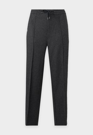 Jeune homme debout portant une chemise bleu marine à manches longues, un pantalon gris foncé à cordon et des baskets blanches sur un fond clair uni.