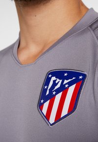 Šedé sportovní tričko s výstřihem do V a krátkými rukávy, které se vyznačuje výrazným znakem se štítovým motivem s červenobílými pruhy a modrými akcenty.