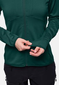 Giacca a zip di un verde scuro con un tessuto strutturato. Design aderente, aperture per il pollice sui polsini e tasche con zip sui lati. Pantaloni neri visibili.