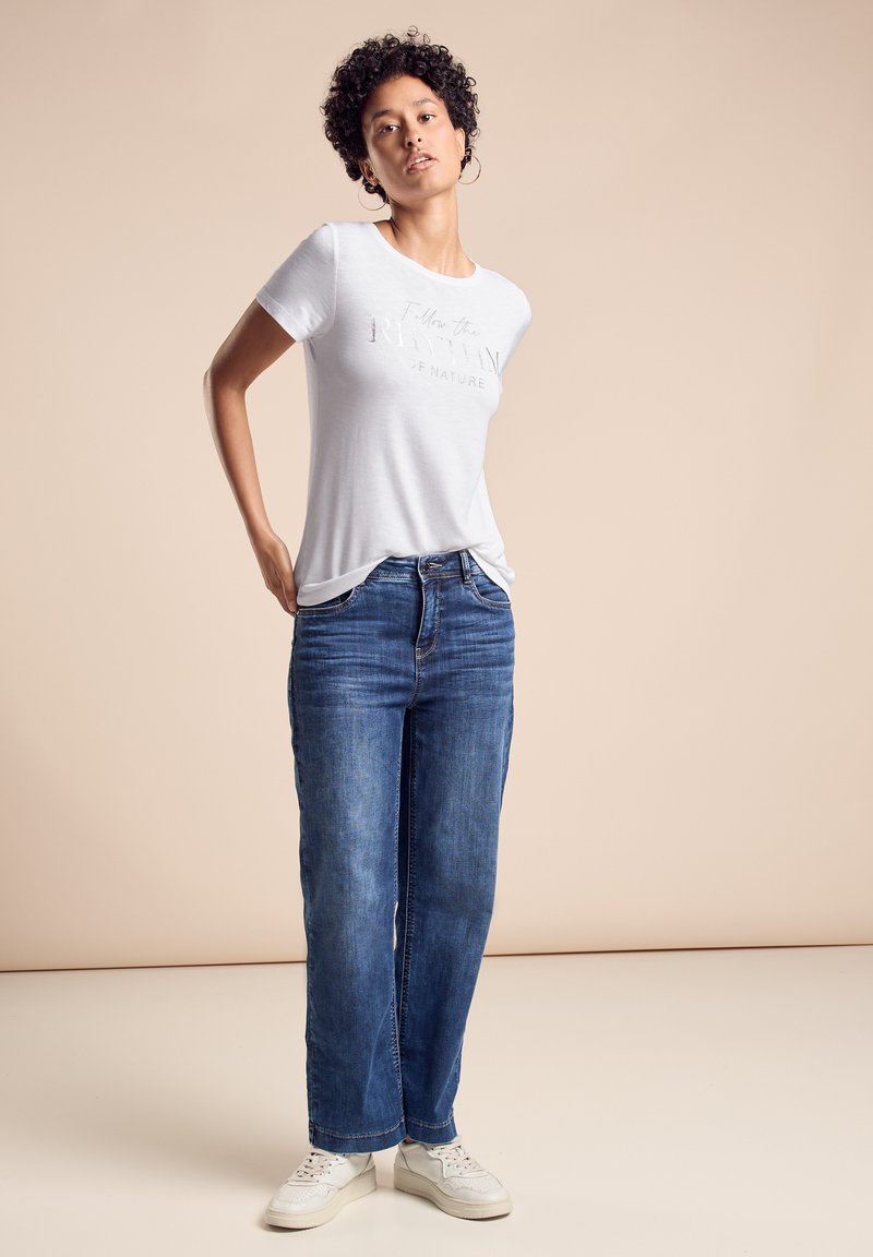 Hvid t-shirt med kortærmer og teksten "Følg naturens rytme" trykt i grå. Båret med blå lige ben jeans og hvide sneakers.