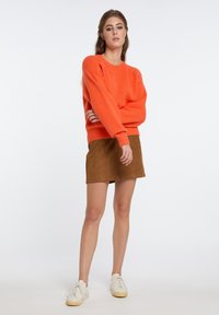Maglione in morbido filato arancione con polsini e fondo a coste, abbinato a una mini gonna in suede marrone. Le sneakers bianche con lacci completano l'outfit.