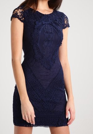 Mujer con un vestido ajustado azul marino de encaje, mangas cortas y patrones intrincados de flores y formas geométricas.