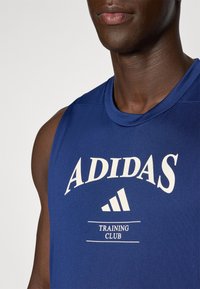 Sininen hihaton urheilupaita, jossa on valkoinen "ADIDAS" -logo ja "TRAINING CLUB" -teksti, sileä tekstiili ja pyöreä kaula-aukko.