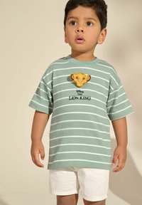 Hele rohelise triibuline T-särk, millel on naeratava Lõvikuningas tegelaskuju plaastriga, koos valgete lühikeste pükste ja lühikeste varrukatega ning mugava lõikega.