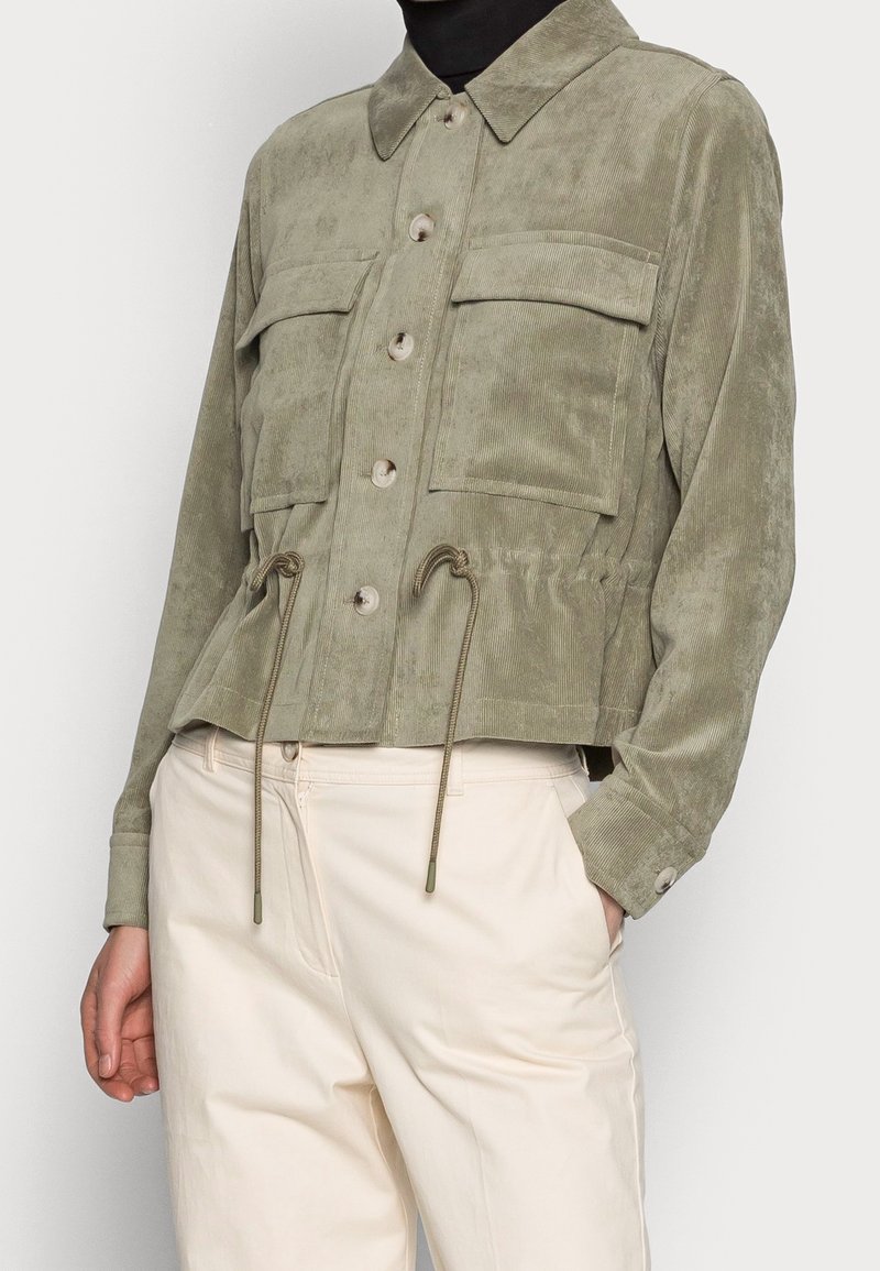 Olive green cropped jacket in soft suede texture with four pockets, button closures, and adjustable drawstring at the waist.