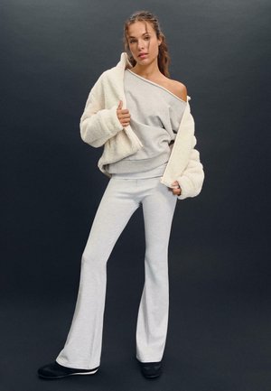 Maglione grigio a maglia e pantaloni svasati coordinati, abbinati a una giacca crema testurizzata. L'outfit presenta una vestibilità rilassata e materiali morbidi.