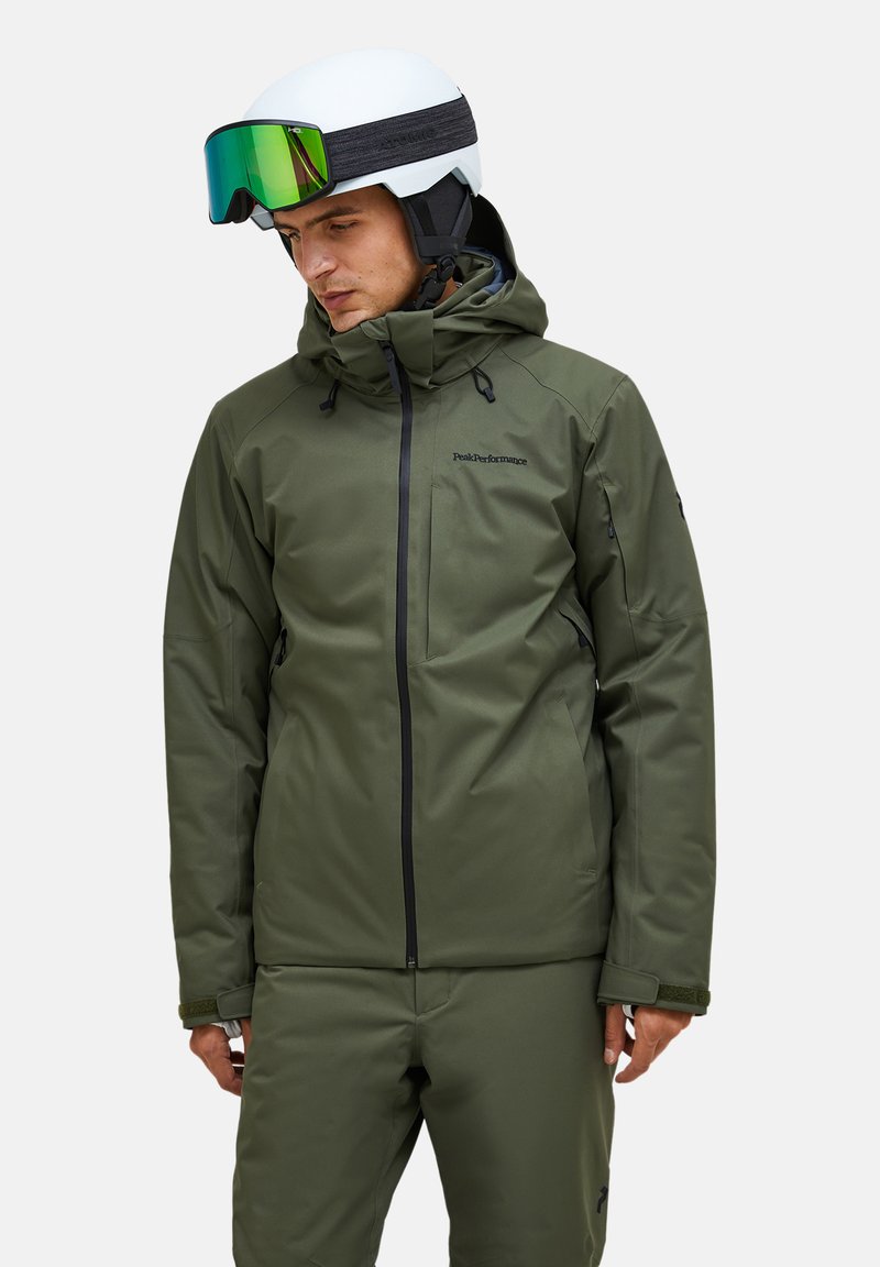 Groene geïsoleerde jas met een rits, verstelbare capuchon en meerdere zakken, gecombineerd met een bijpassende broek en een witte helm met een vizier.